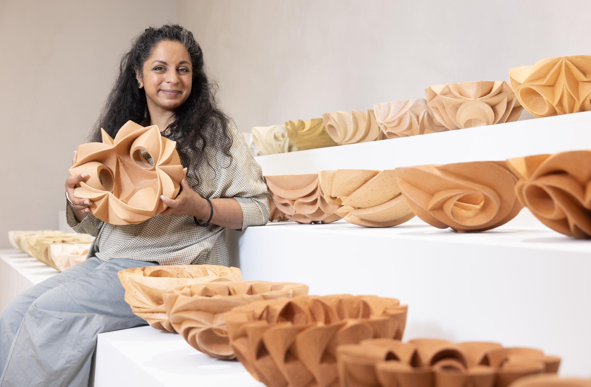 Halima Cassell poses with an intricately carved ceramic piece, surrounded by her sculptural bowls on display.