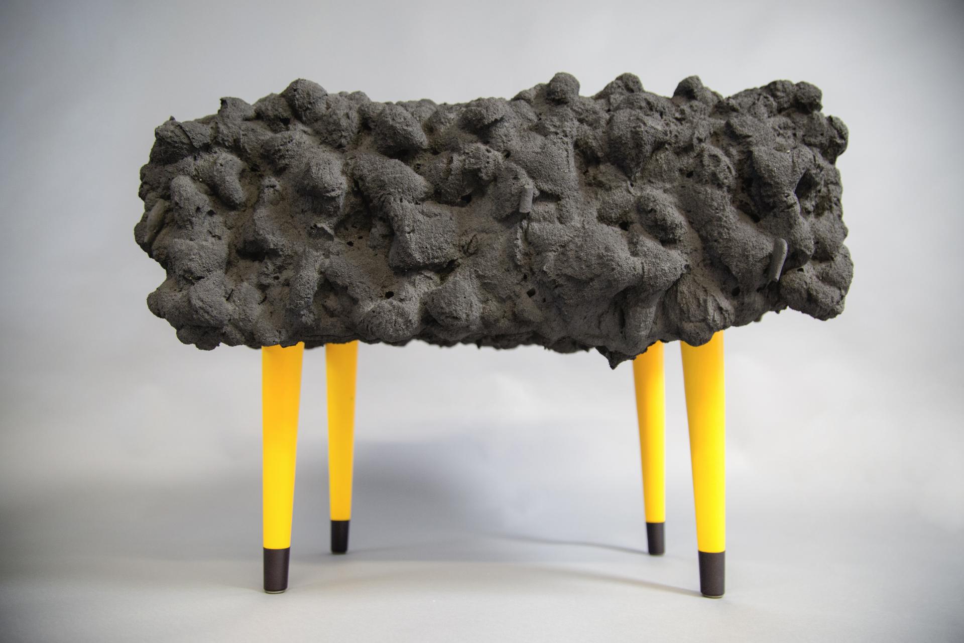 Contemporary furniture piece with a textured black surface resembling erosion, supported by vibrant yellow legs, representing nature's influence.