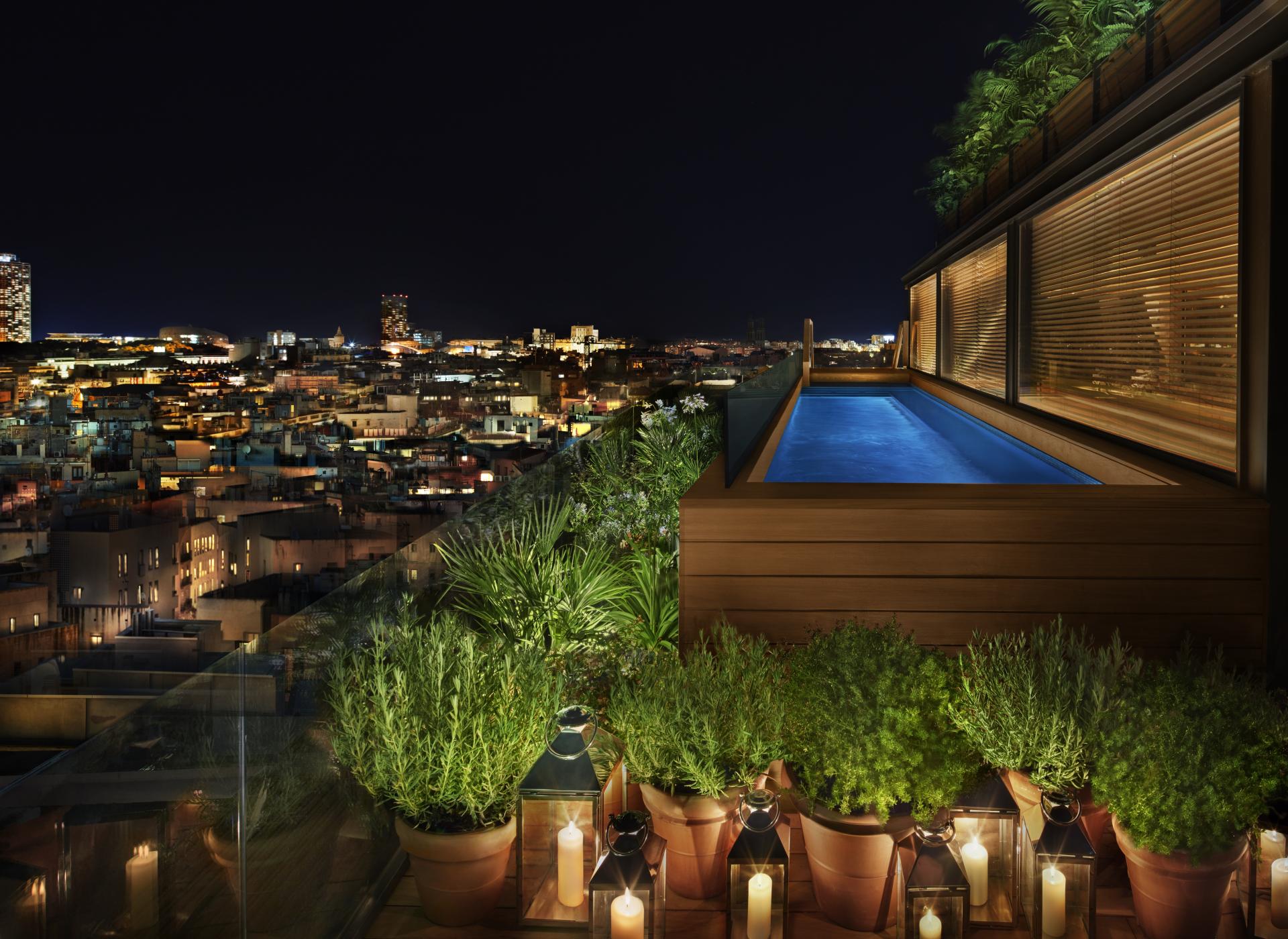 Luxurious rooftop pool surrounded by greenery, overlooking the illuminated skyline of Barcelona at night.