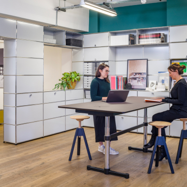 Two women collaborate at a modern table in the USM Clerkenwell showroom, surrounded by modular furniture and storage solutions.