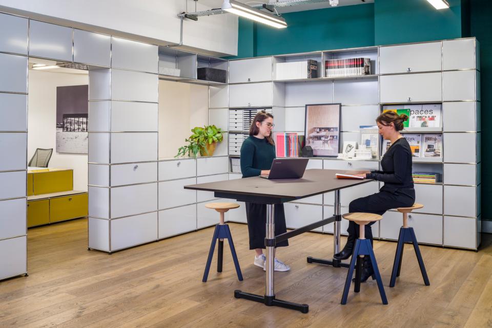 Two women collaborate at a modern table in the USM Clerkenwell showroom, surrounded by modular furniture and storage solutions.