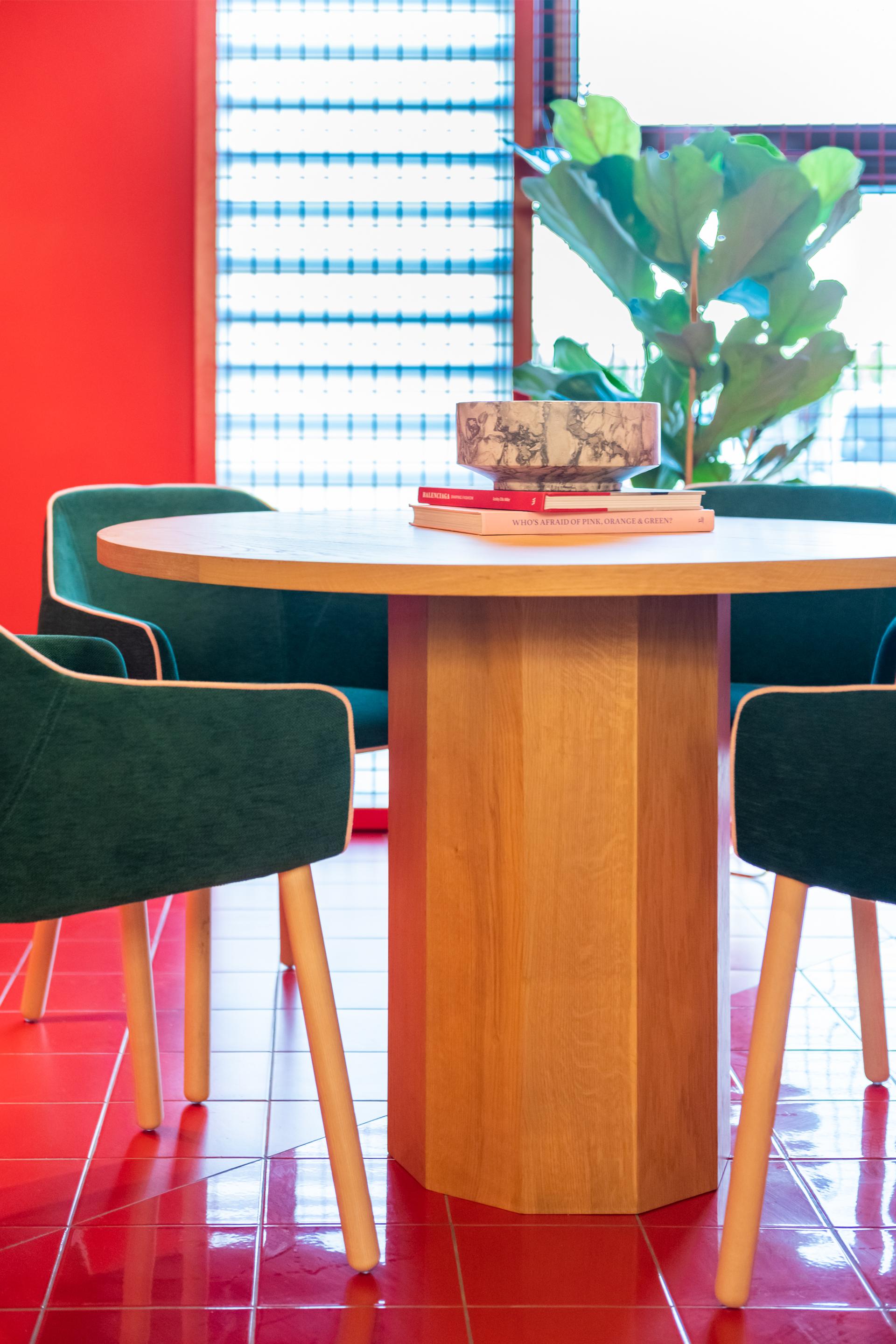 Modern dining area featuring a round wooden table, green chairs, art books, and a decorative bowl against a vibrant backdrop.