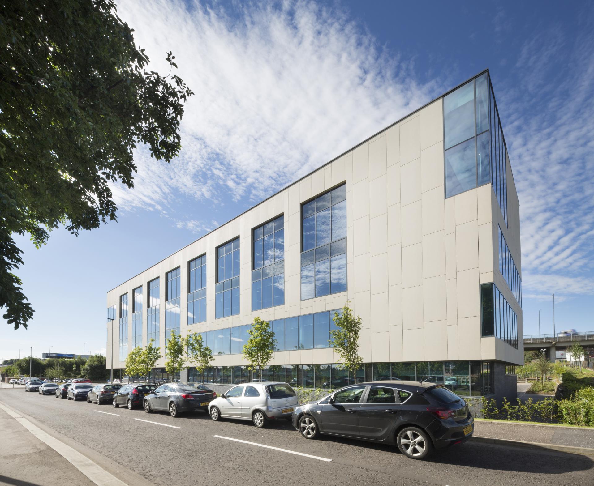 Modern glass-fronted building in Glasgow, surrounded by green trees and parked cars, reflecting urban sustainability efforts.