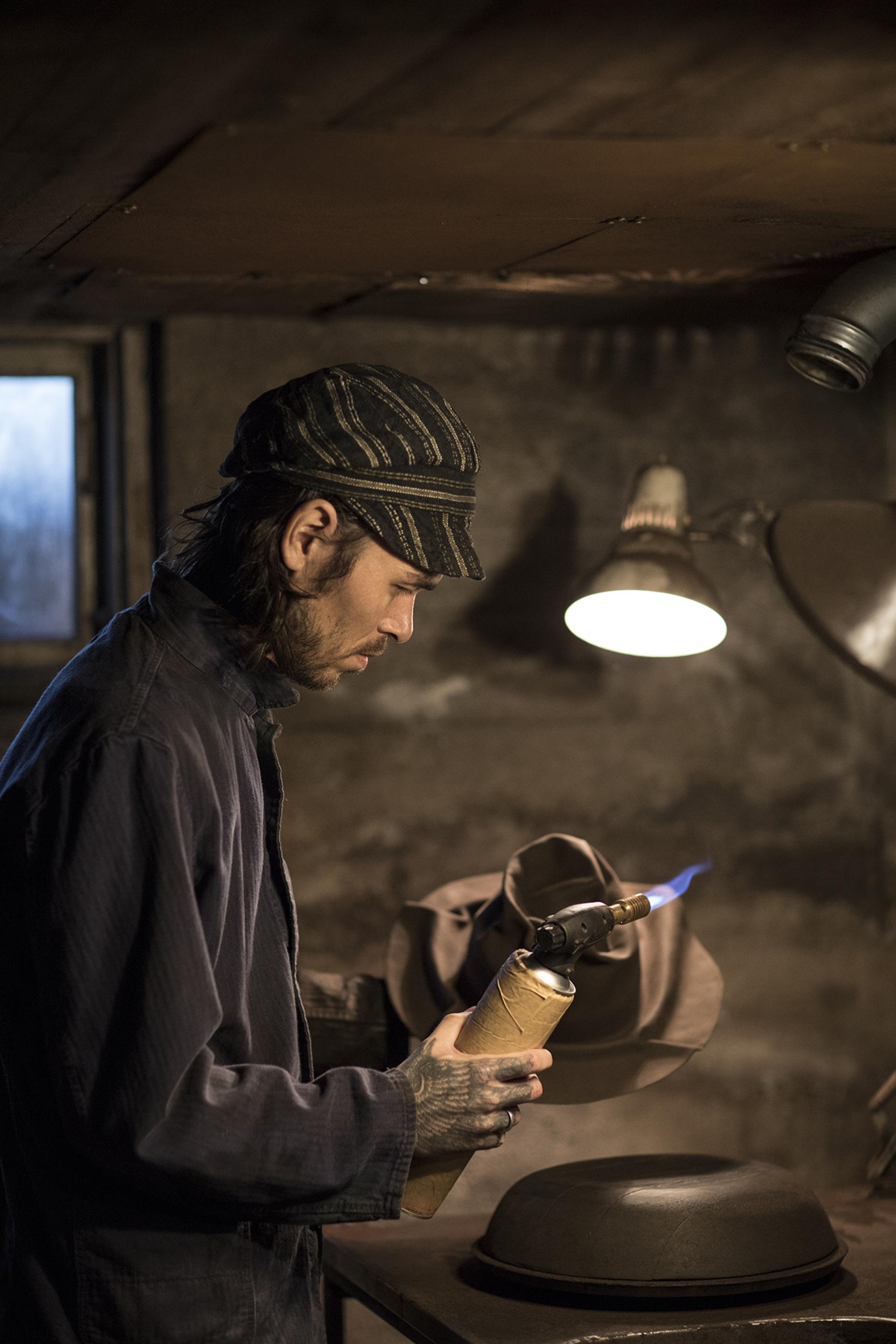 Craftsman using a torch in Horisaki's workshop, creating traditional hats surrounded by Småland’s scenic forest atmosphere.