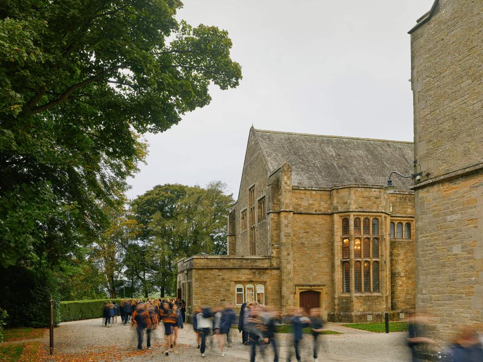 Students in smart uniforms walk past the historic Queens Hall at Sedbergh School, framed by lush greenery.