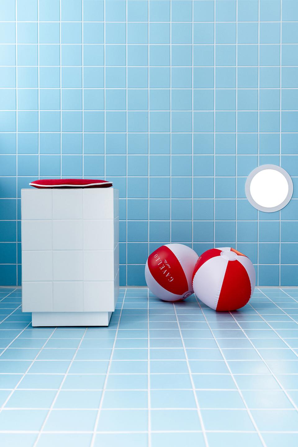 Bright blue tiled space featuring a white pedestal and red-and-white beach balls, evoking a playful poolside atmosphere.