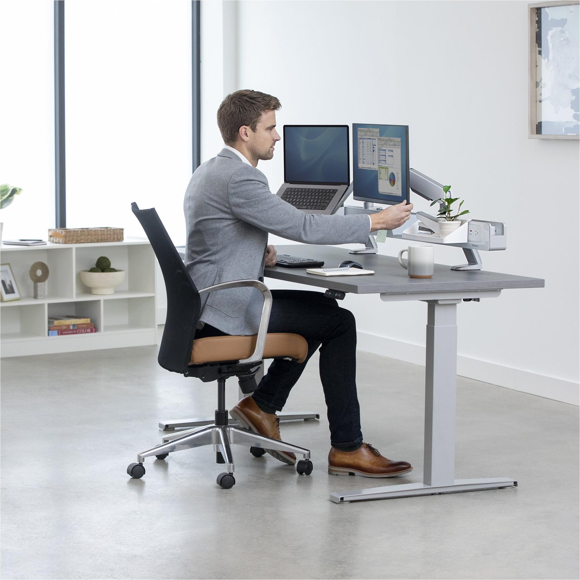 Modern ergonomic workspace featuring a man at a height-adjustable desk with dual monitors and a plant.