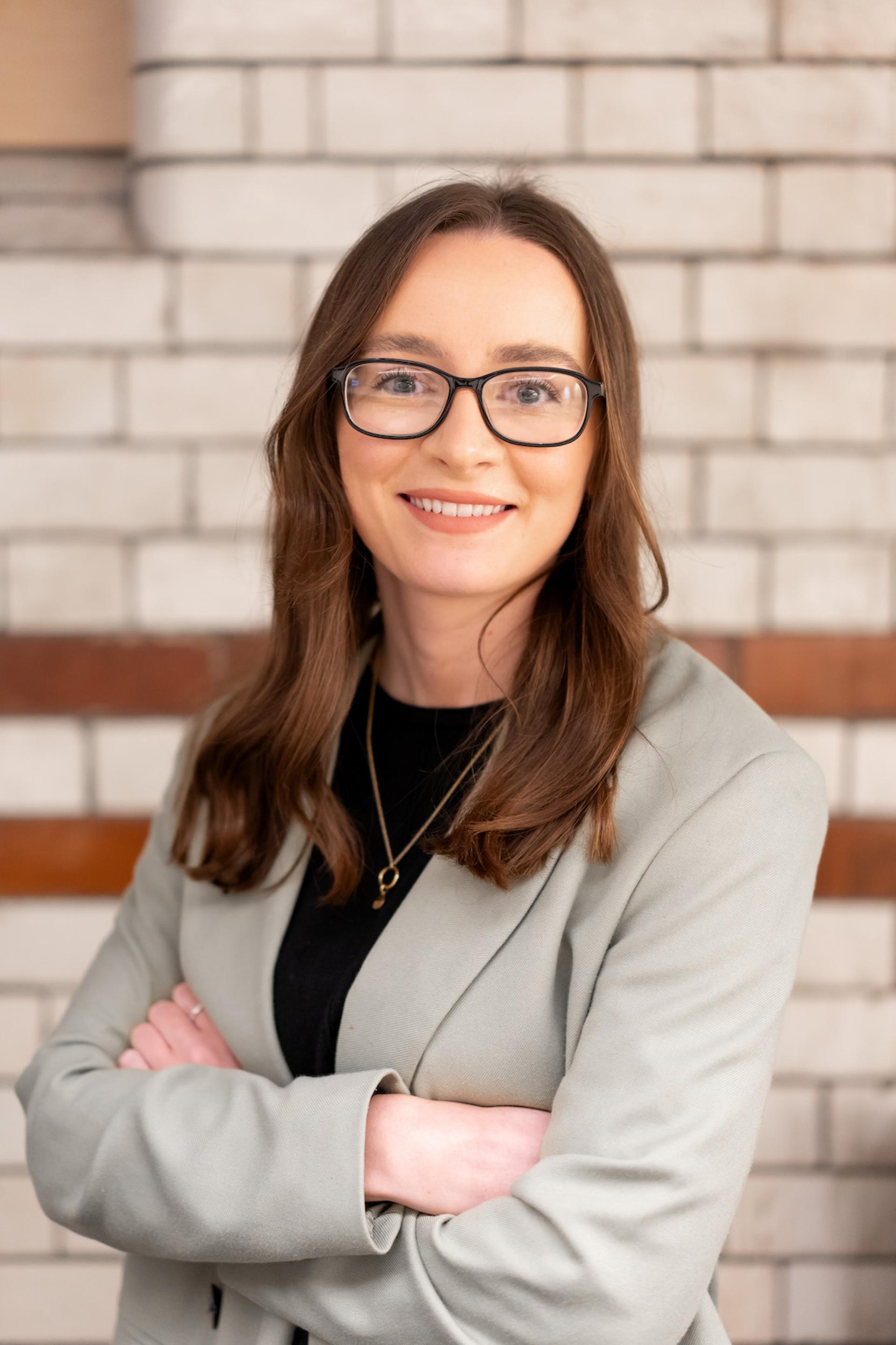 Professional woman with long hair wearing a blazer, smiling confidently against a modern brick background, highlighting sustainability discussions.