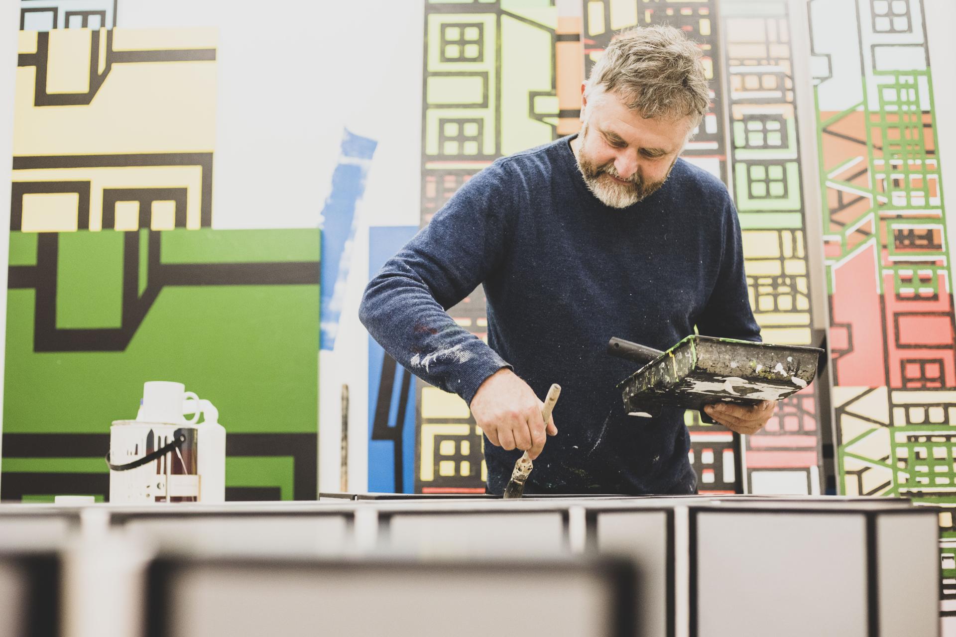 Richard Woods painting creatively in his studio, surrounded by vibrant abstract designs and showcasing his passion for raw materials.