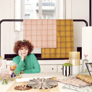 Serena Confalonieri poses creatively among vibrant textiles and unique glassware, showcasing her innovative design style and artistic vision.