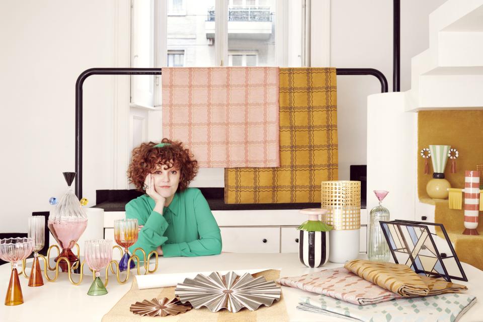 Serena Confalonieri poses creatively among vibrant textiles and unique glassware, showcasing her innovative design style and artistic vision.