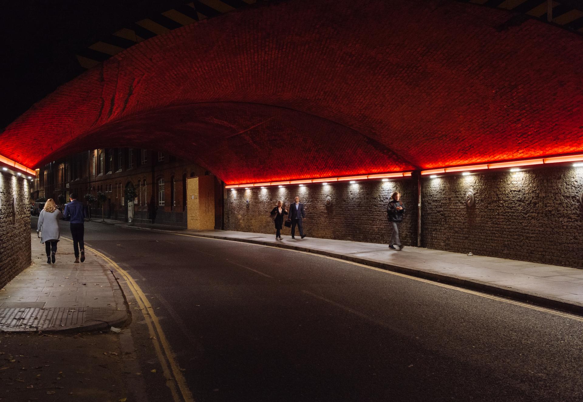 Illuminated urban walkway under a red brick arch, featuring pedestrians and modern street lighting at night.