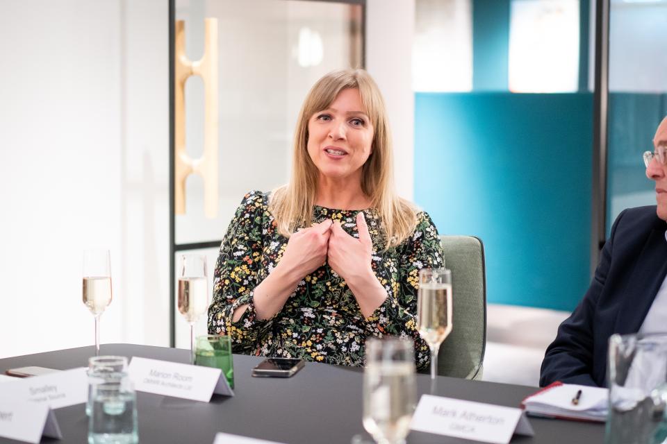A woman in a floral dress discusses sustainability during a seminar, surrounded by participants at a table.