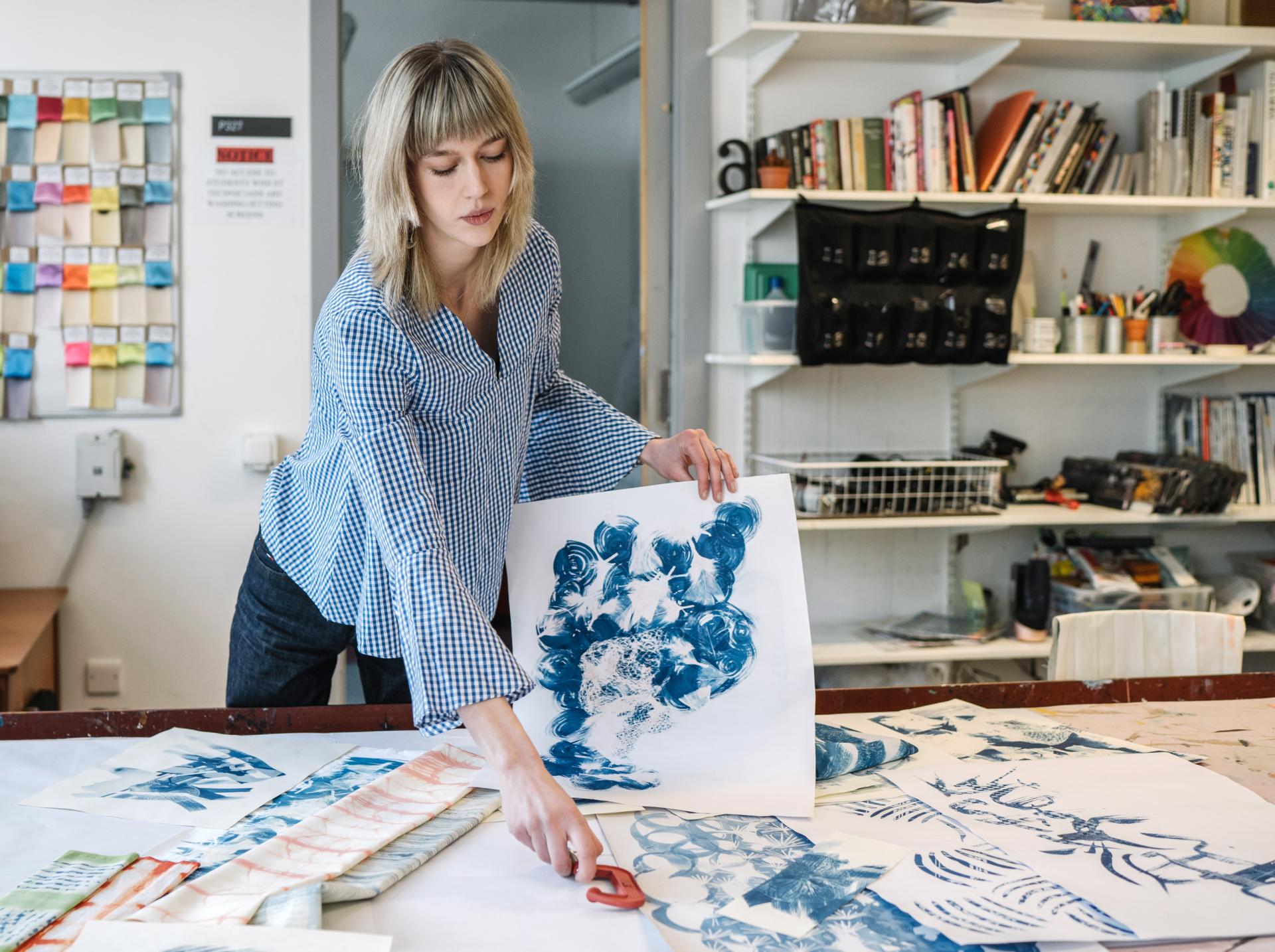 A designer arranging blue-patterned prints on a workspace filled with various textile samples and materials.