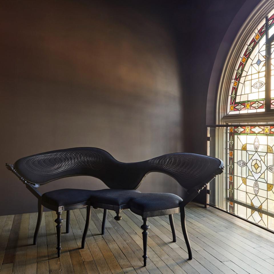 Elegant black sculptural sofa with ornate detailing, positioned near a stunning stained glass window in San Francisco's Carpenters Workshop Gallery.