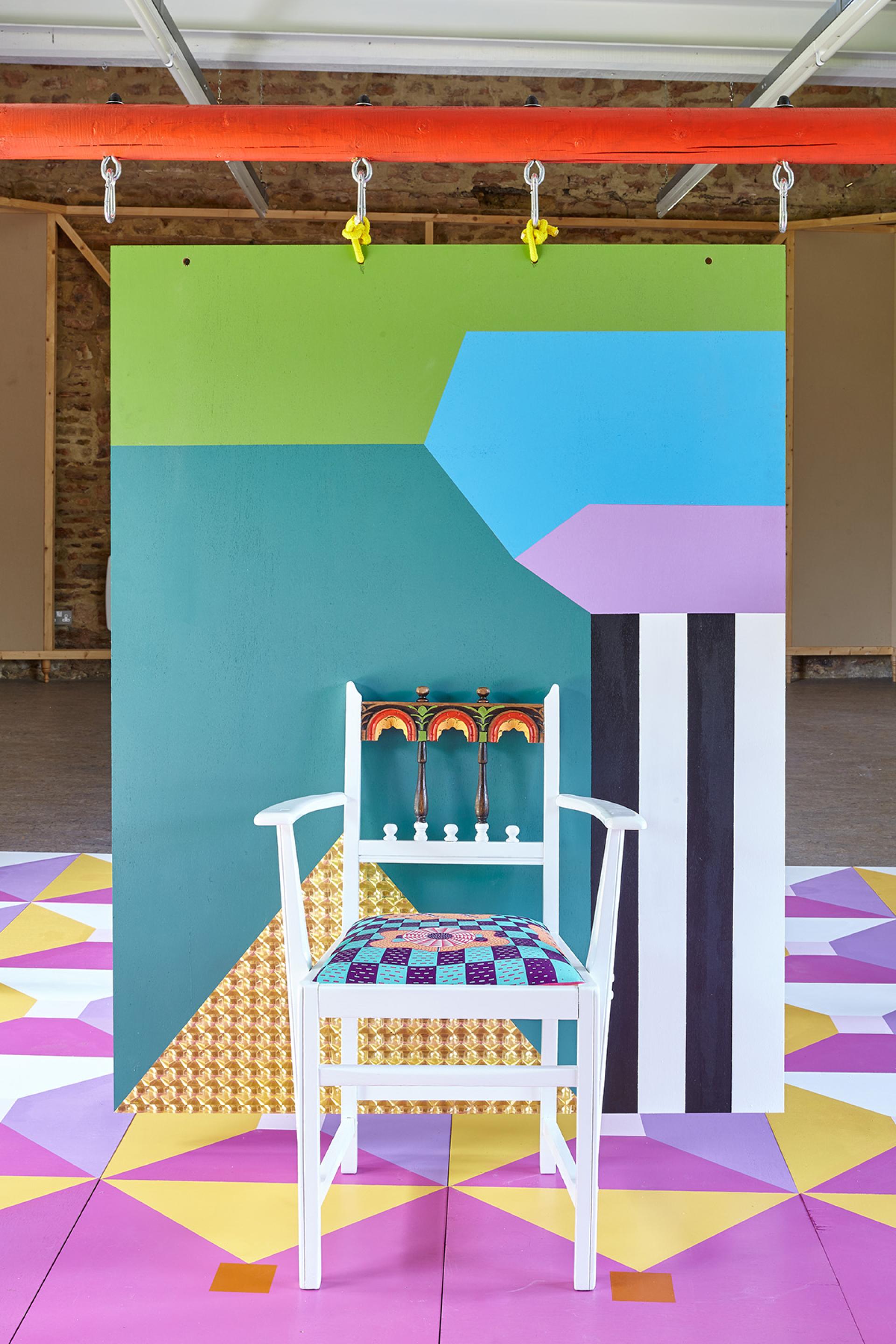 Colorful artistically designed chair in front of a vibrant mural, showcasing Yinka Ilori's unique style and passion for upcycled furniture.