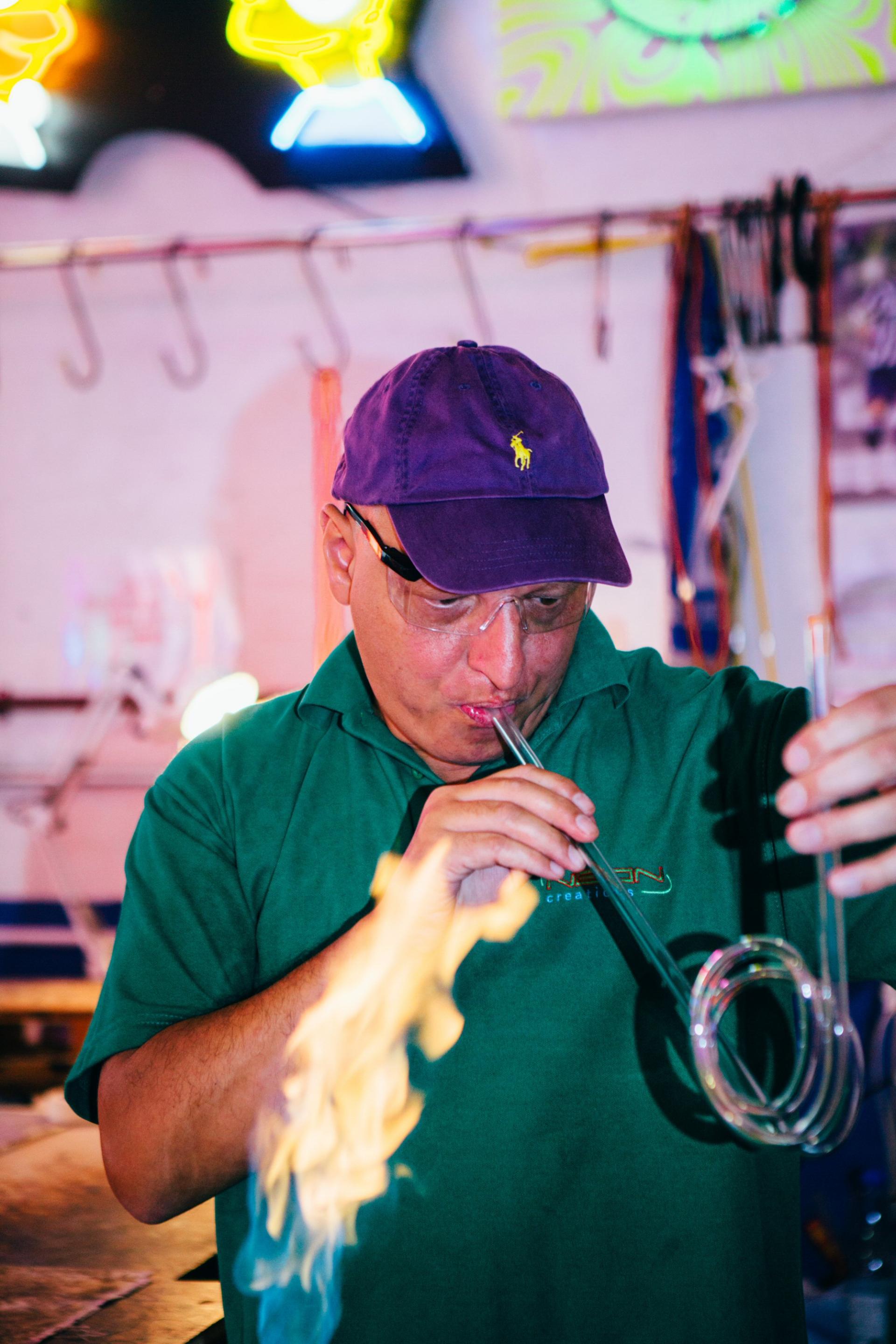 Craftsman shaping glass tubing while blowing flames, creating neon signs in a vibrant workshop setting.
