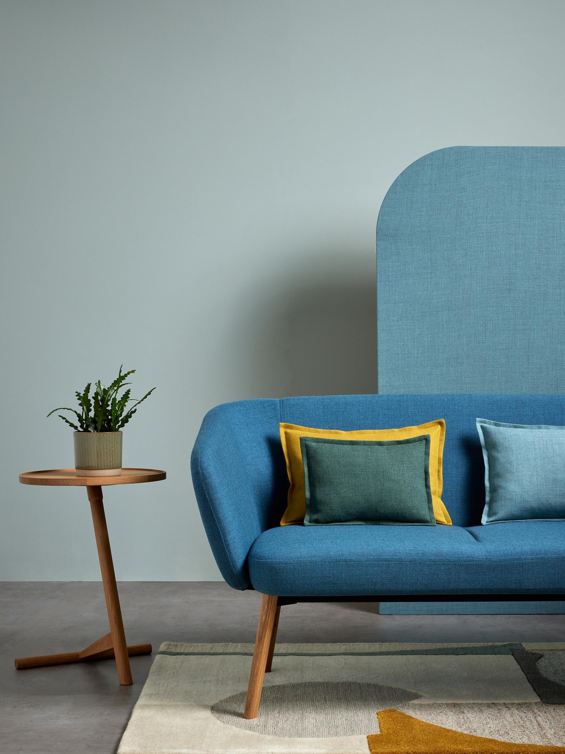 Modern blue sofa with colorful cushions, a wooden side table, and decorative plant, showcasing Camira's Sumi fabric palette.