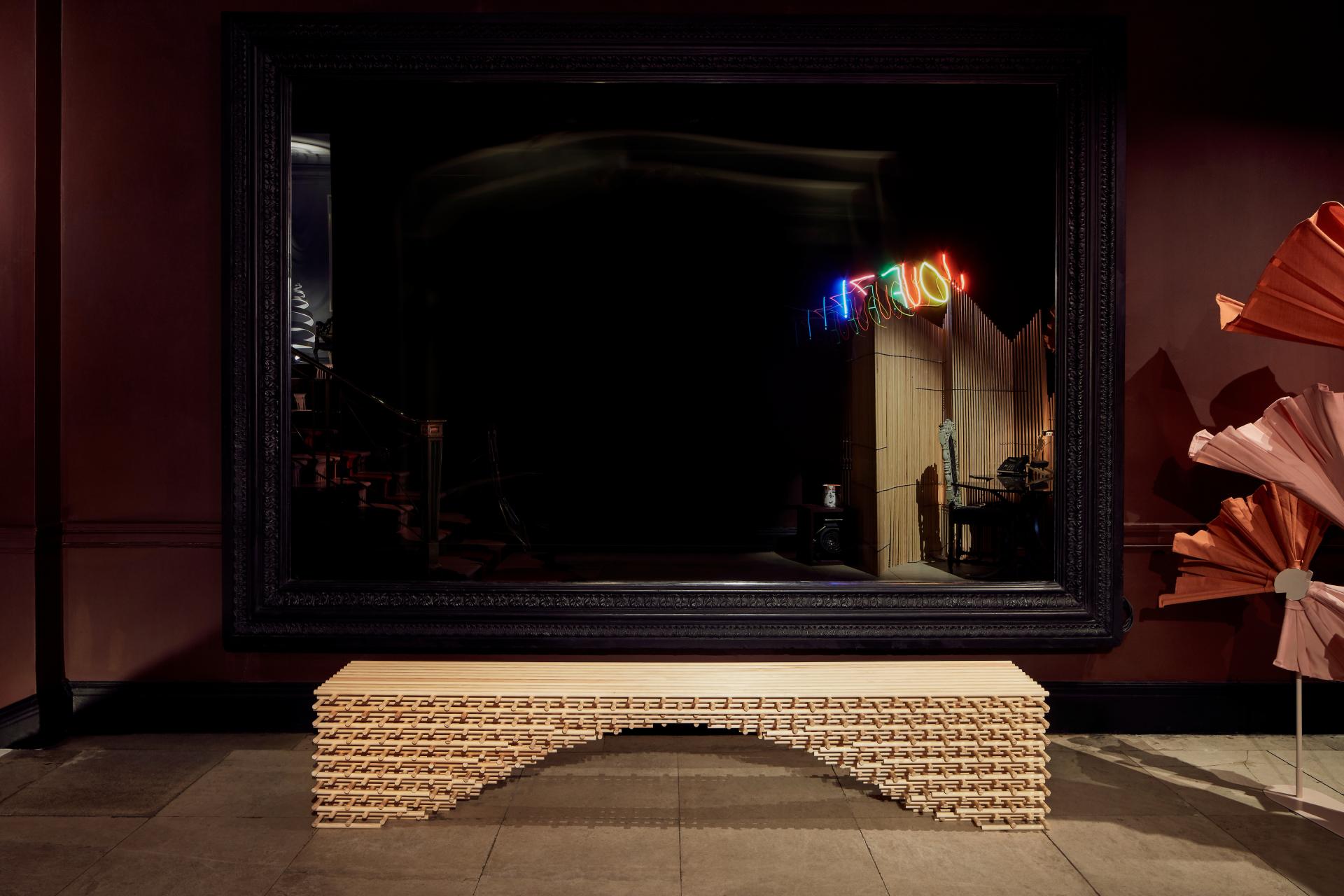 Sculptural wooden bench beneath a large ornate mirror, reflecting colorful neon lights at the A Second Life exhibition.