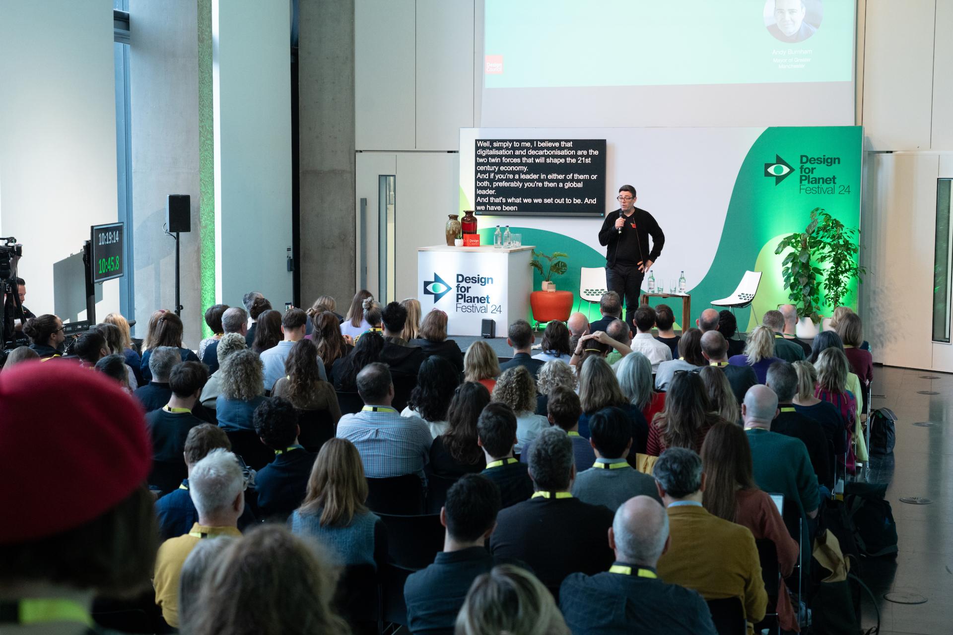 A speaker presents ideas on sustainability at the Design for Planet Festival, engaging an audience in a modern venue.