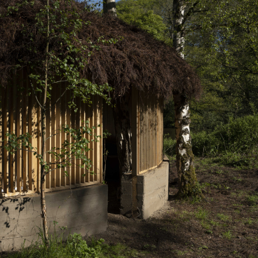 A sustainable art installation featuring a wooden structure with a natural thatched roof surrounded by lush greenery.
