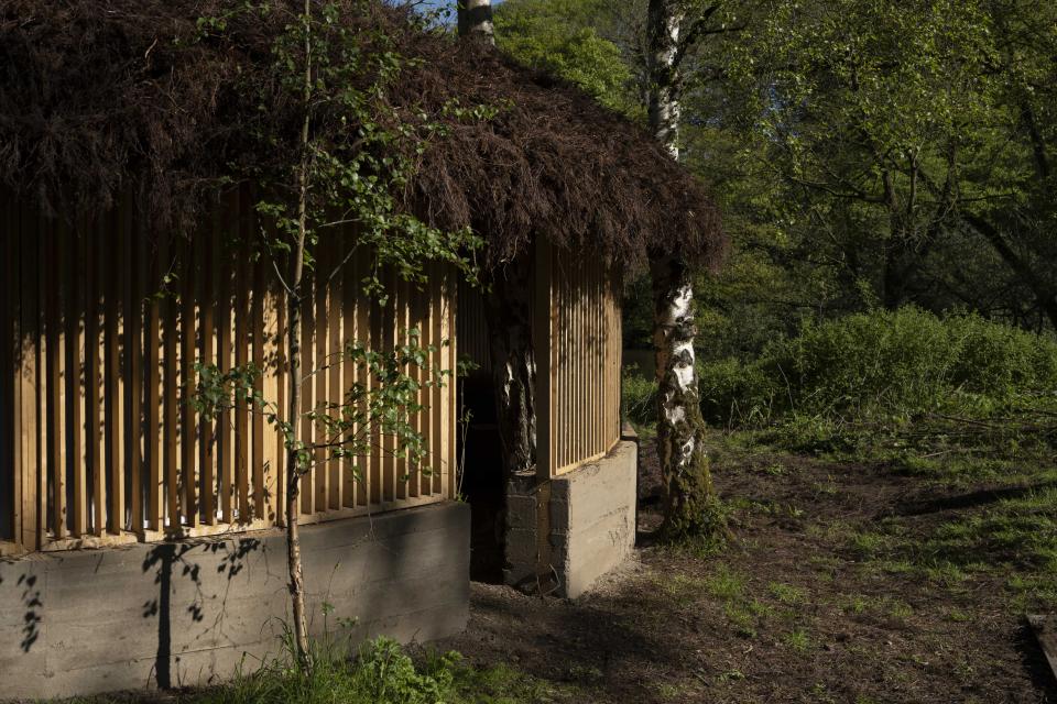 A sustainable art installation featuring a wooden structure with a natural thatched roof surrounded by lush greenery.