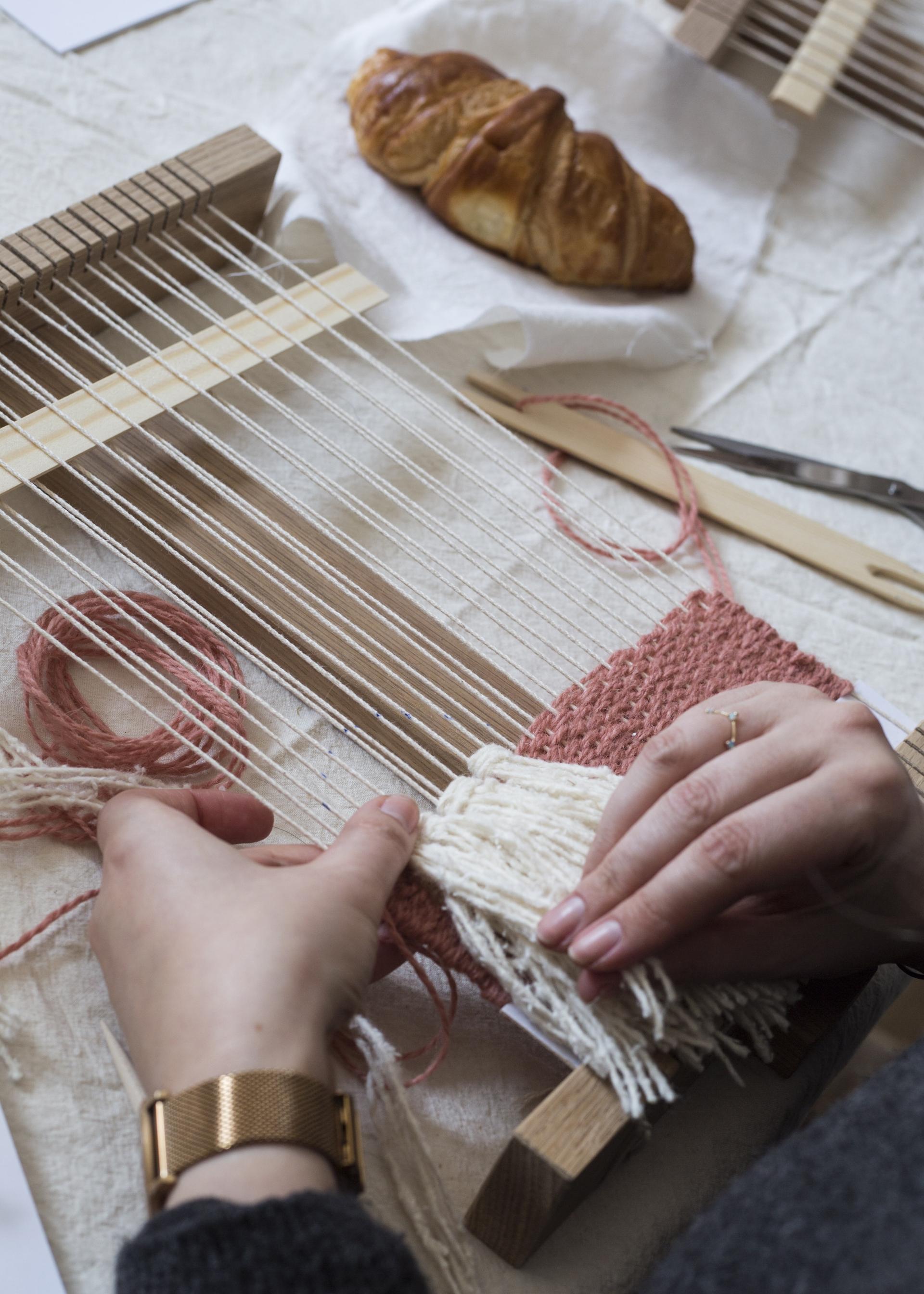 Hands weaving tapestry with pink and white yarn on a wooden loom, accompanied by a croissant on the table.