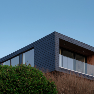 Modern coastal home featuring CUPACLAD slate cladding, large windows, and wooden balcony, surrounded by greenery.