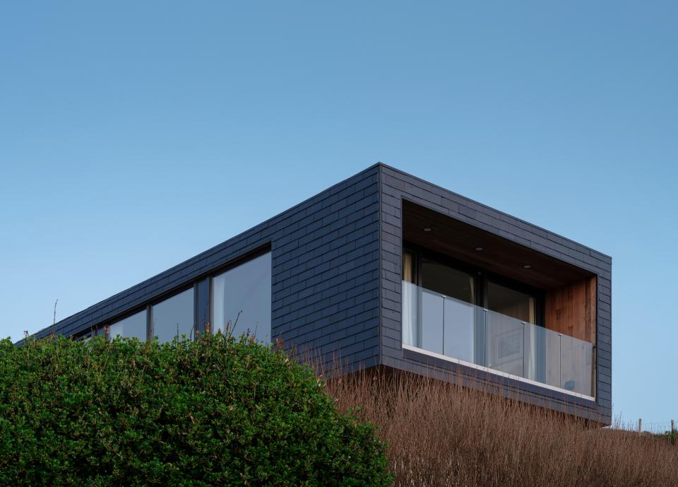 Modern coastal home featuring CUPACLAD slate cladding, large windows, and wooden balcony, surrounded by greenery.