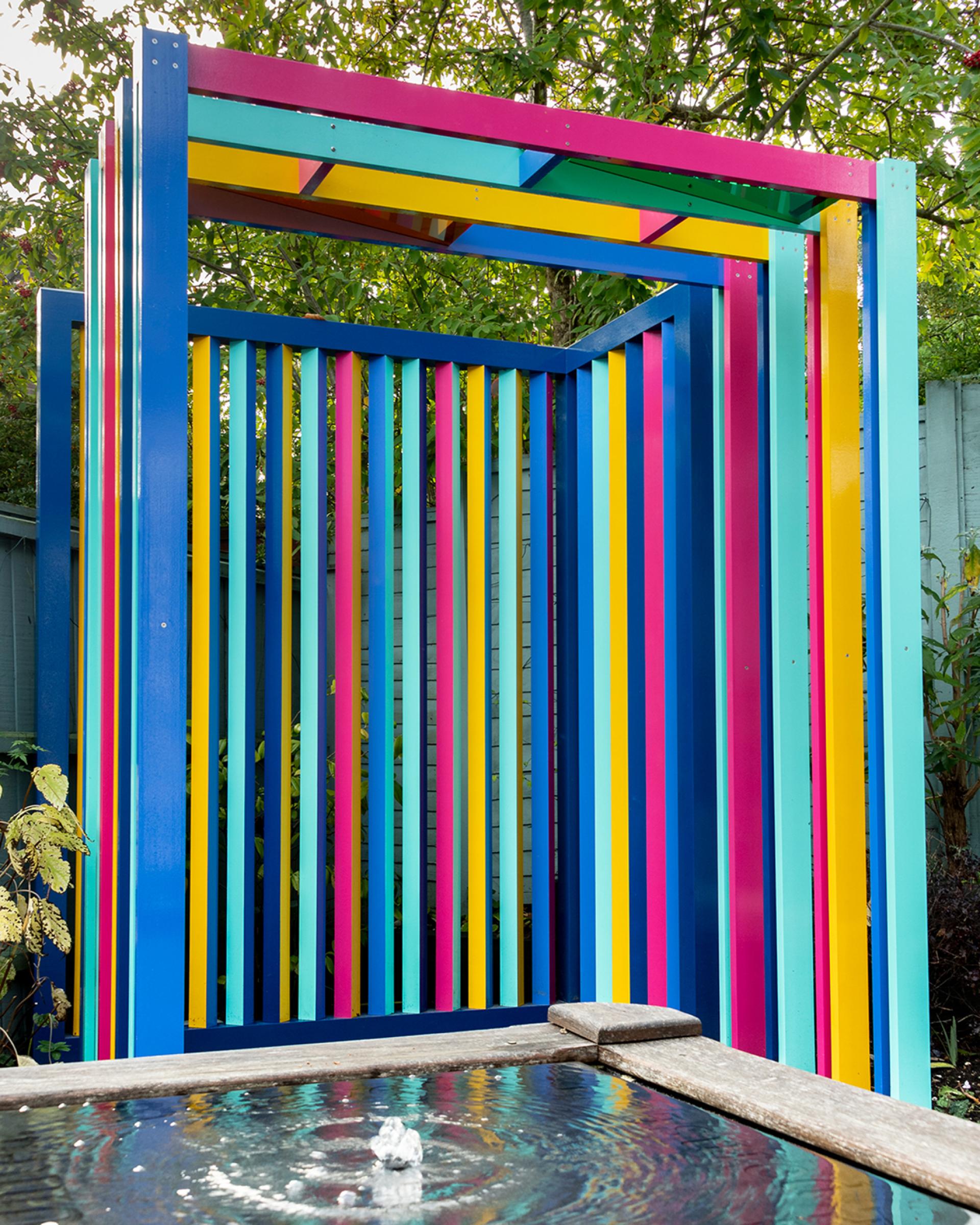 Colorful modern outdoor structure with vertical slats, complementing a serene water feature in a garden setting.