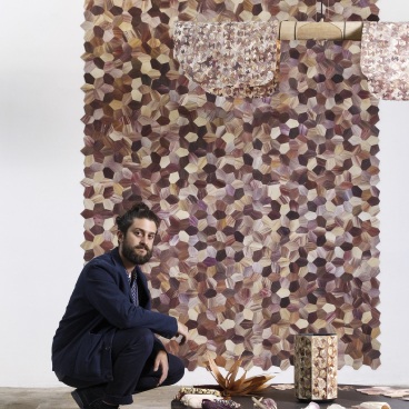 A man kneels beside a table displaying Totomoxtle objects and a colorful hexagonal wall installation.