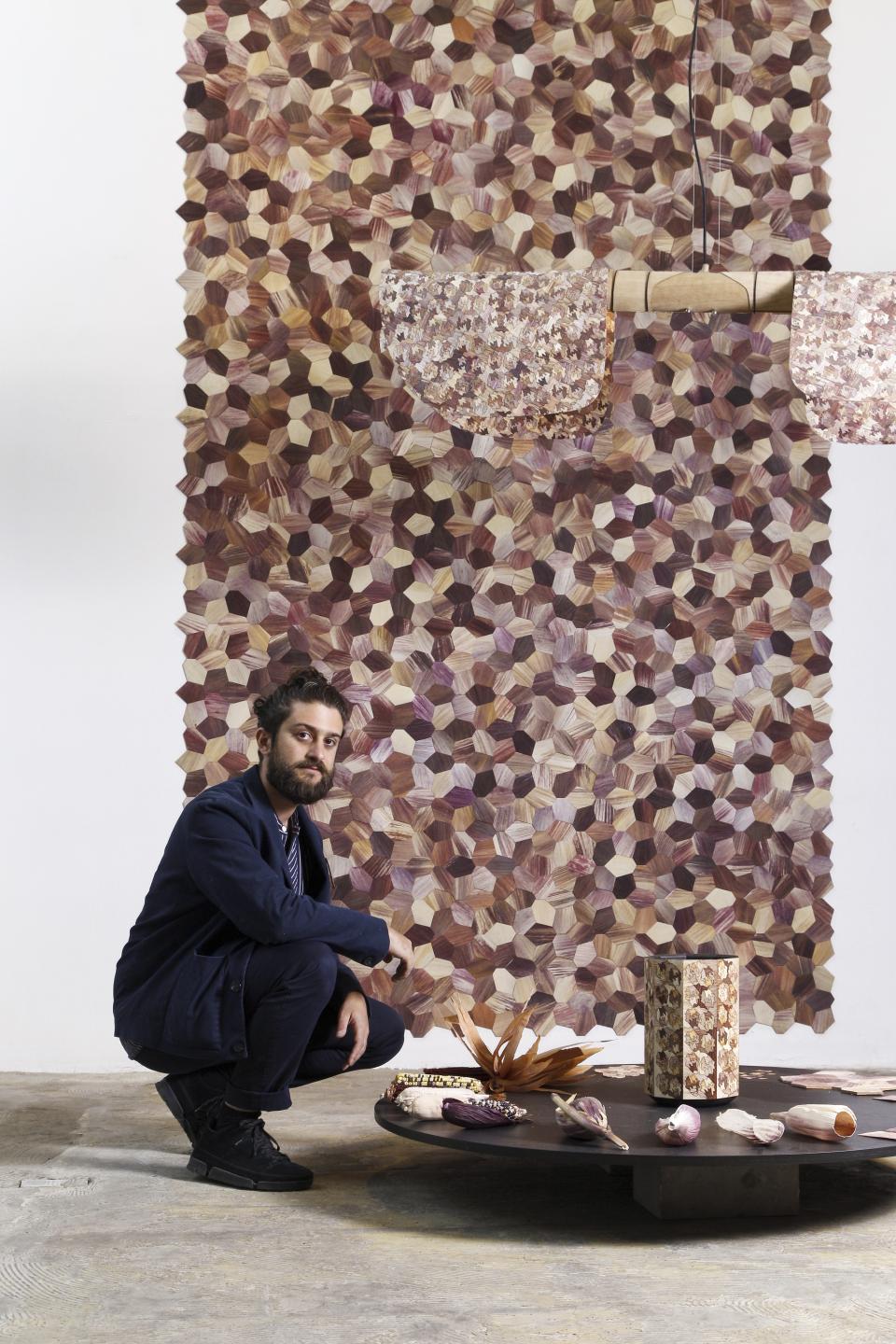 A man kneels beside a table displaying Totomoxtle objects and a colorful hexagonal wall installation.