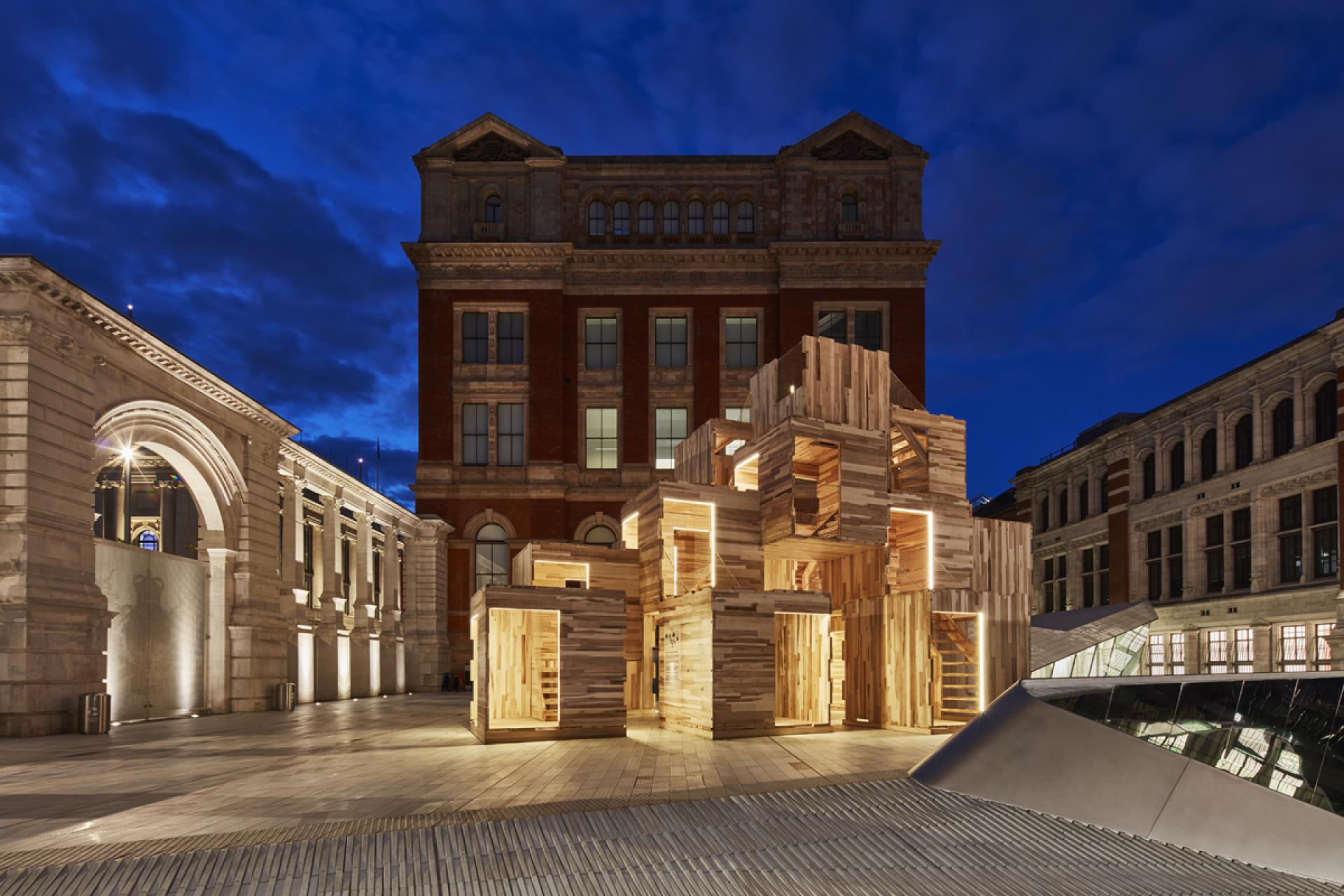 MultiPly wooden pavilion illuminated at night, showcasing carbon-neutral architecture amidst historic buildings.