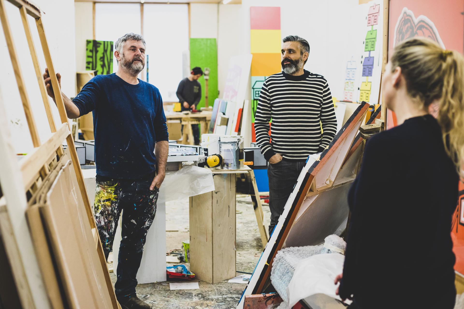 Artists engaged in conversation in a vibrant studio filled with colorful materials and sculptures, celebrating creativity and discarded art.