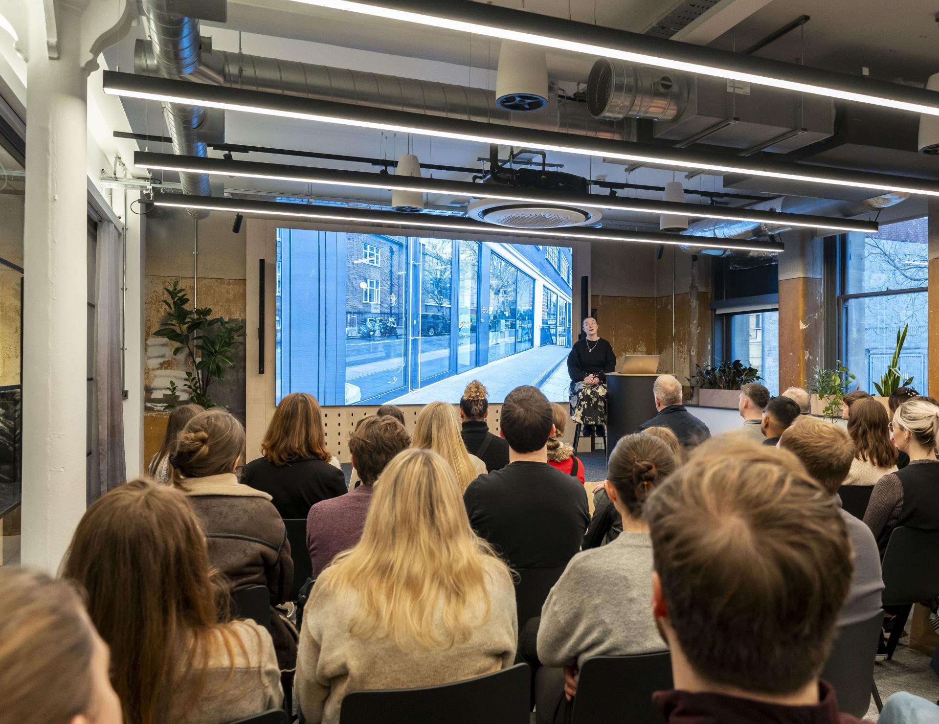 Architect discussing neurodiversity and inclusive design to an engaged audience in a modern setting with large windows.
