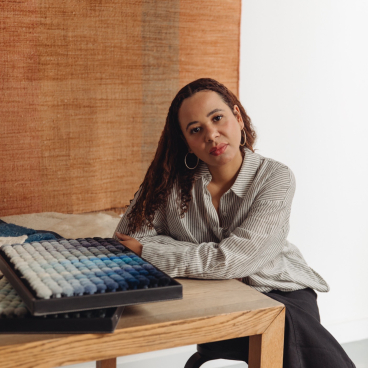 Dharma Taylor poses thoughtfully beside an intricately textured textile artwork, emphasizing themes of memory and narrative.