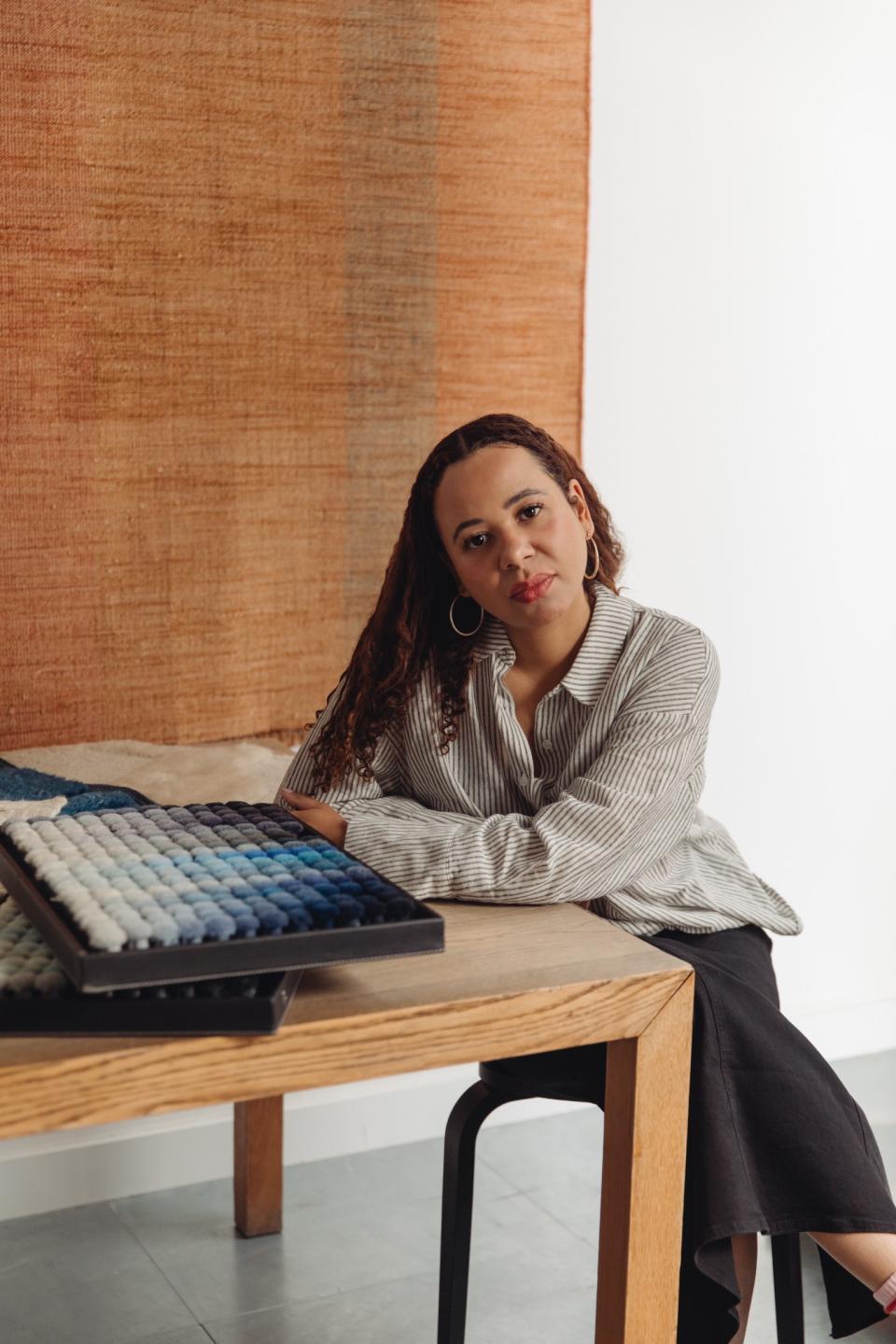 Dharma Taylor poses thoughtfully beside an intricately textured textile artwork, emphasizing themes of memory and narrative.