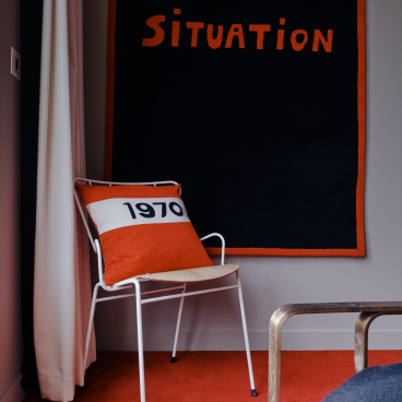 Elegant interior featuring a white chair with an orange cushion, against a bold black wall tapestry saying "Situation."