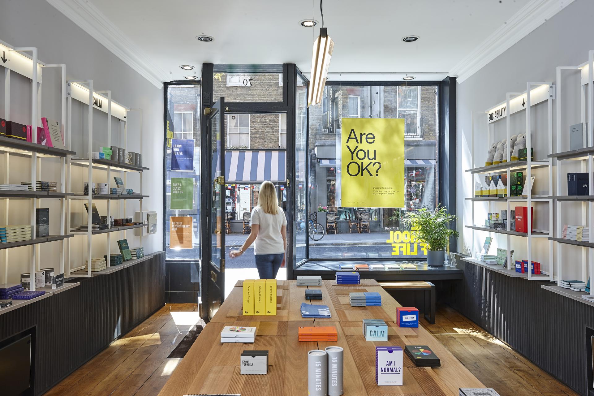 Brightly lit interior of a design studio featuring a wooden table with colorful books and a cheerful "Are You OK?" poster.