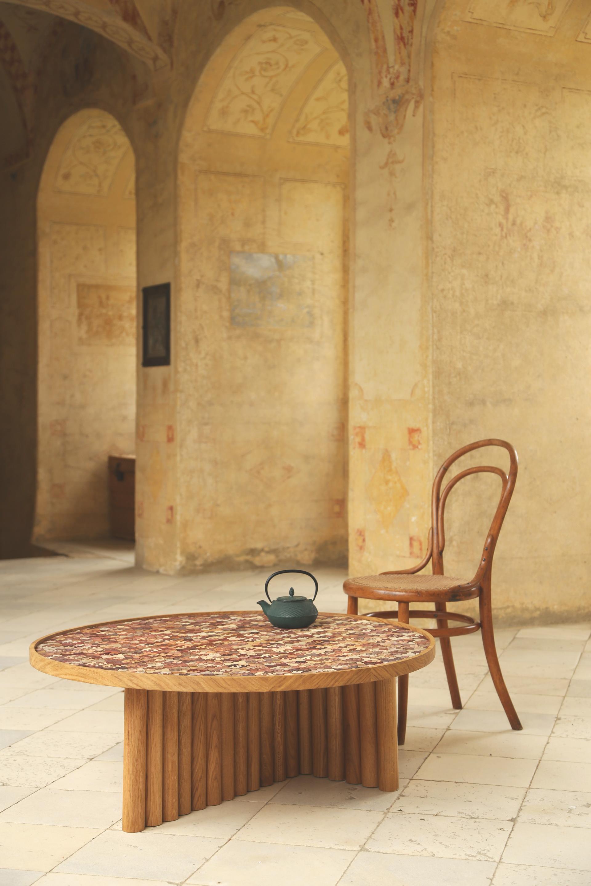 A circular wooden table featuring a colorful Totomoxtle mosaic top, accompanied by a teapot and a vintage chair.
