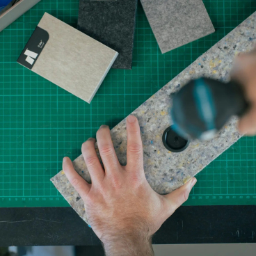 Hand holding a textured acoustic panel while drilling, surrounded by felt samples on a workspace, highlighting Autex Acoustics' sustainable design.