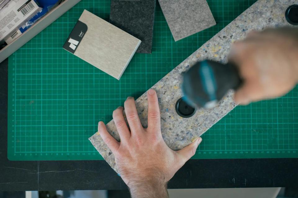 Hand holding a textured acoustic panel while drilling, surrounded by felt samples on a workspace, highlighting Autex Acoustics' sustainable design.