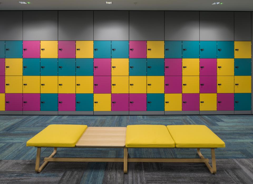 Brightly colored lockers in varying hues of blue, pink, and yellow complement a modern seating area in a WELL-certified office.