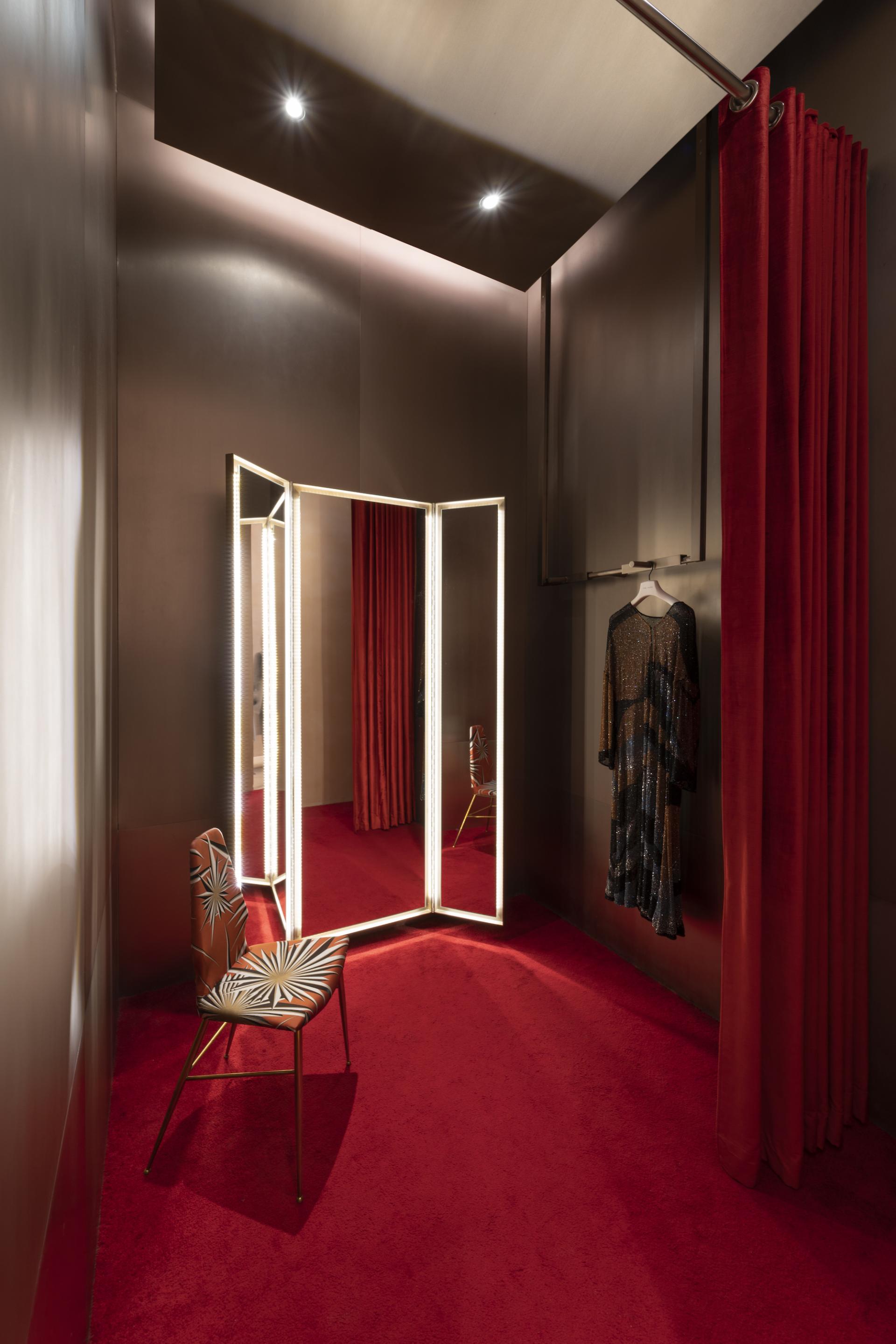 Elegant fitting room featuring a stylish black dress, vibrant red curtains, and a decorative chair with a unique design.