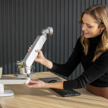 A woman adjusts a Rising™ monitor arm on a desk, showcasing its adaptable design for inclusive workspaces.