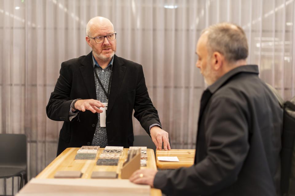 Two men discuss design materials at a wooden table, celebrating creativity at Material Source Studio Manchester's anniversary event.