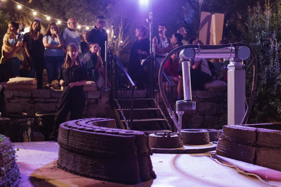 3D printer creating a building structure from clay, with an audience observing at Jerusalem Design Week 2022.
