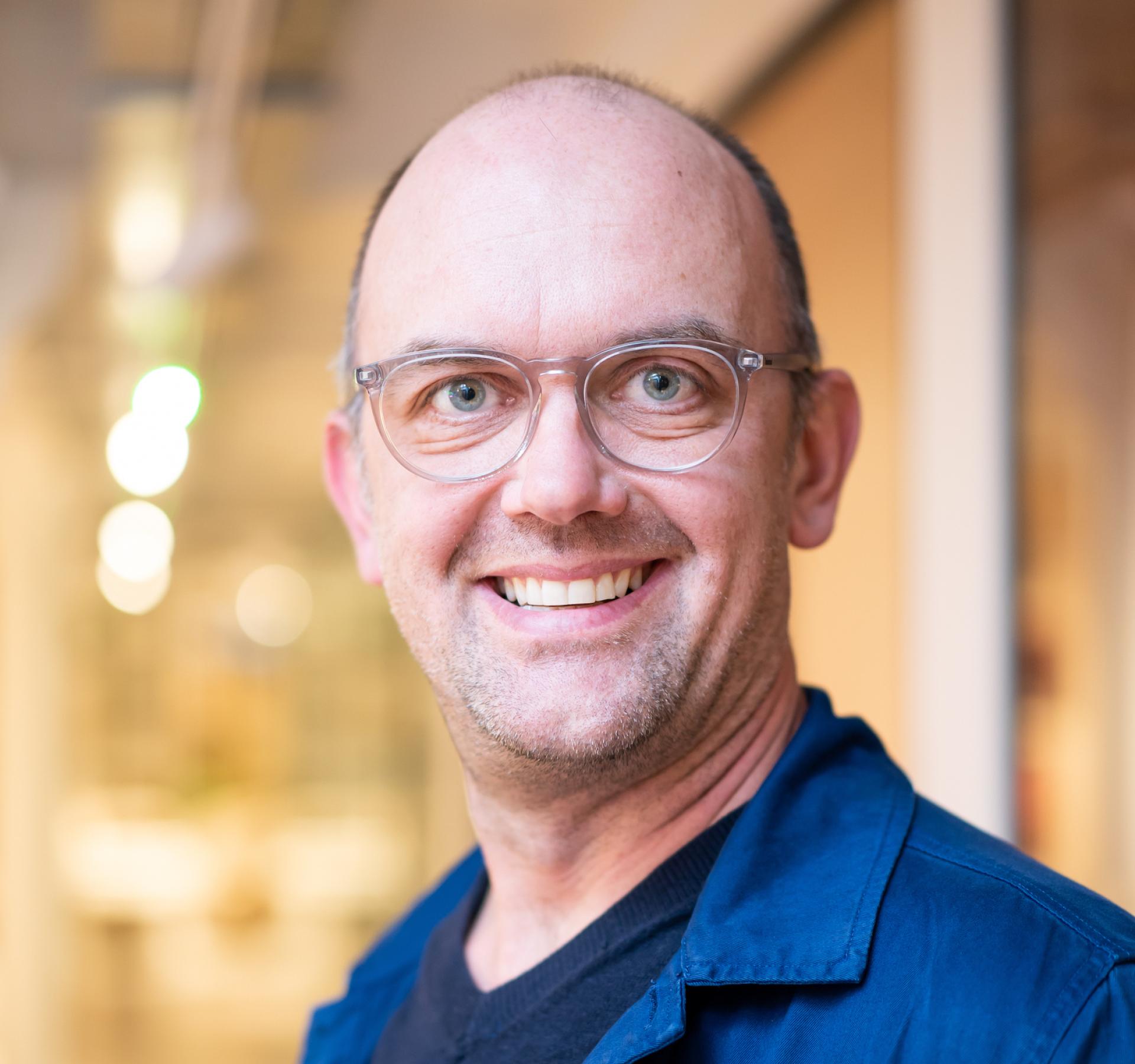 Smiling educator in glasses, wearing a blue jacket, reflecting on the future of education in a modern learning environment.