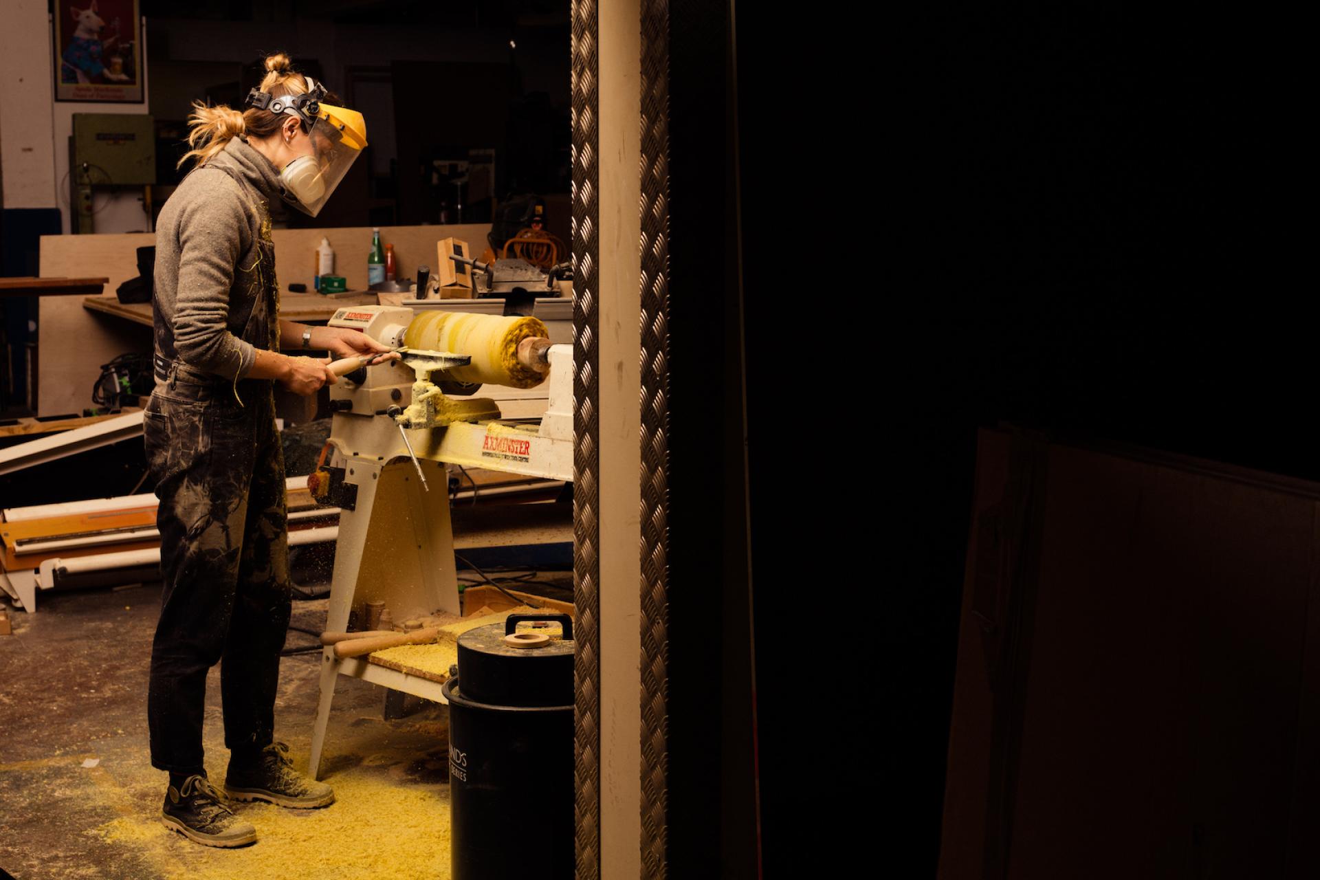 Monika Swindells working on a wood lathe in her creative studio, surrounded by shavings and tools.