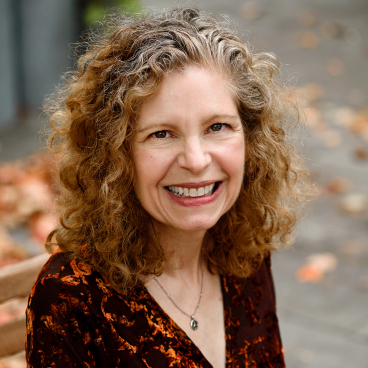 Smiling woman with curly hair, wearing a patterned top, seated outdoors surrounded by autumn leaves, discussing workplace futurism.